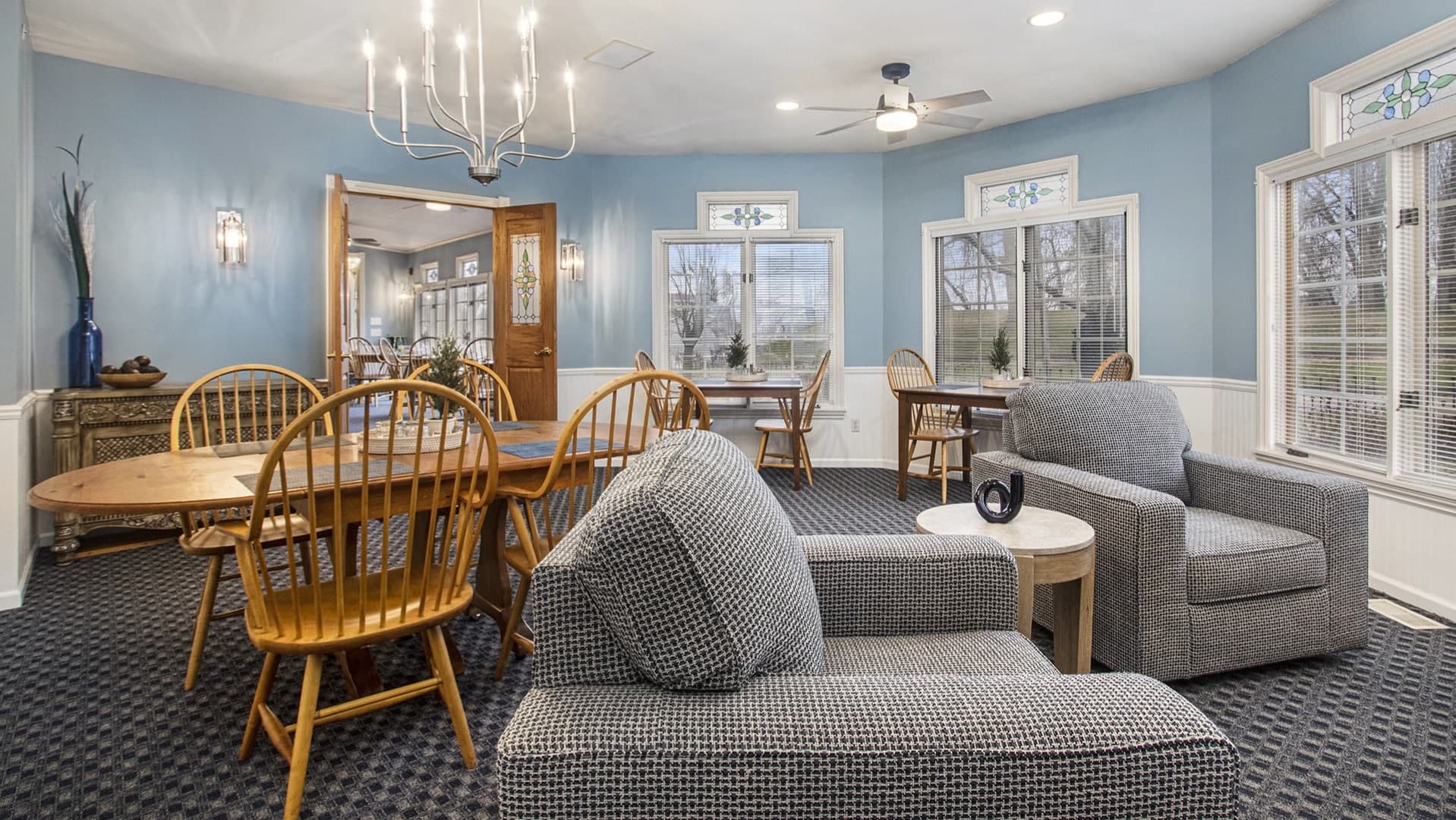 A cozy dining area features wooden tables, chairs, and a comfortable seating arrangement against a light blue wall.