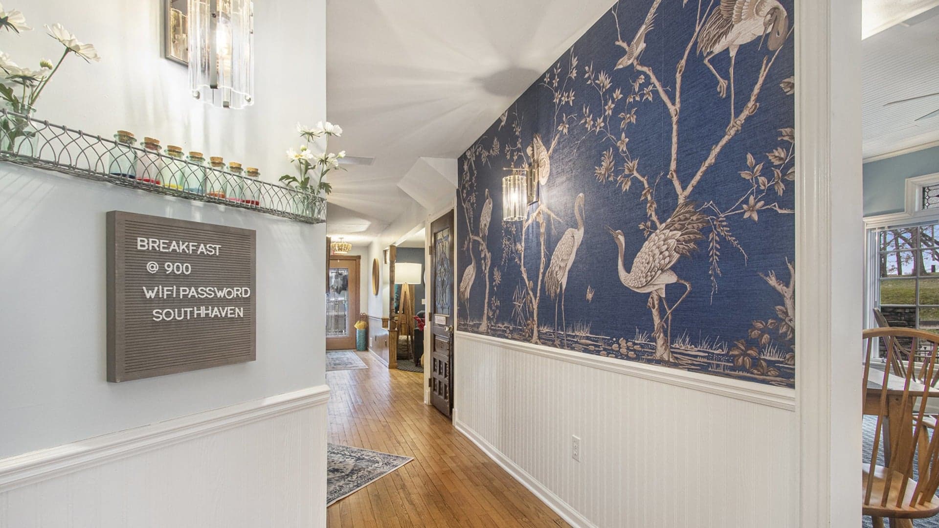 A hallway featuring a decorative blue wallpaper with cranes, a sign for breakfast details, and lit sconces.
