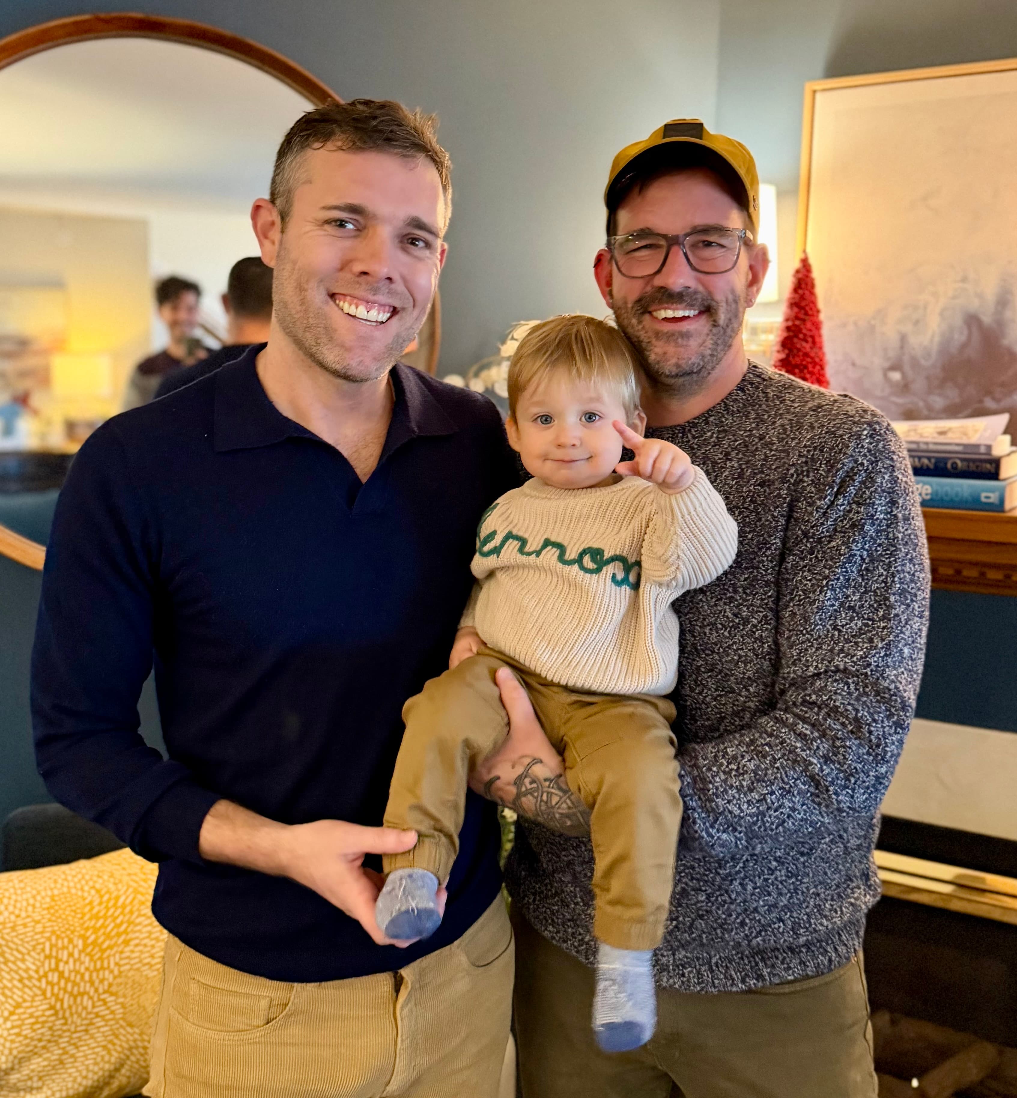 Two men smile while holding a toddler in a cozy living room setting.