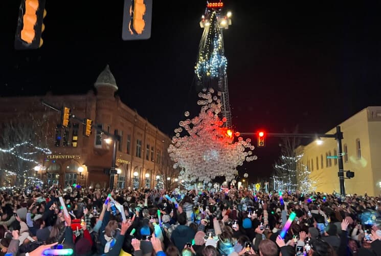 A large crowd celebrates at night while a glowing ball descends from a tall structure amidst festive lights.