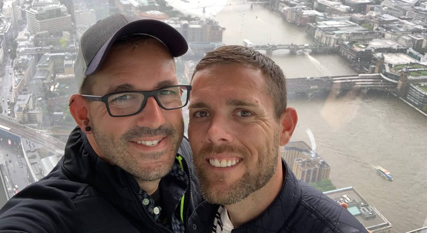 Two men take a smiling selfie with a cityscape view in the background.