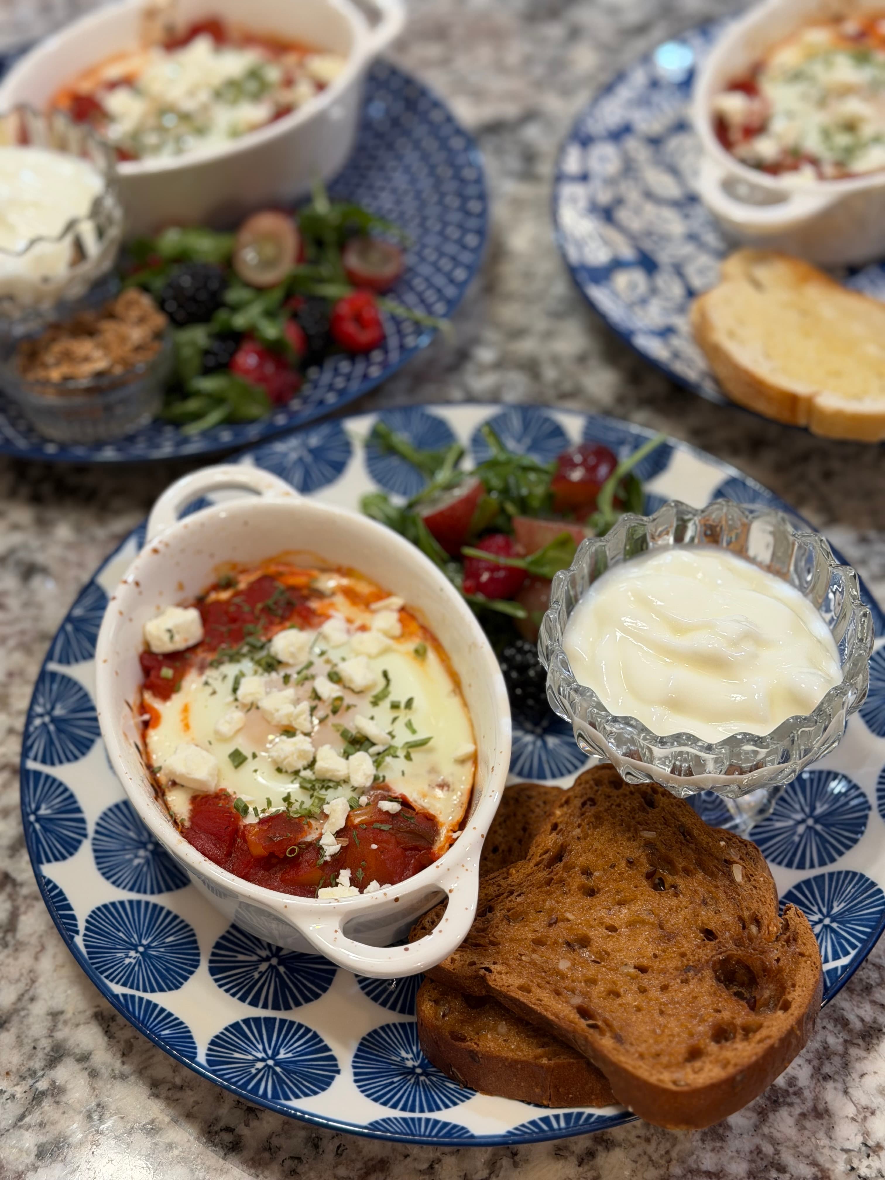 A plate featuring baked eggs with tomato sauce and feta, accompanied by yogurt, toast, and a side of mixed berries and greens.