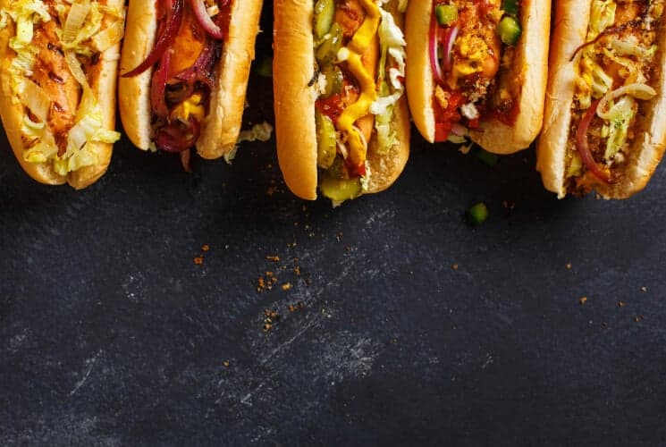 A row of colorful hot dogs topped with various garnishes on a dark surface.