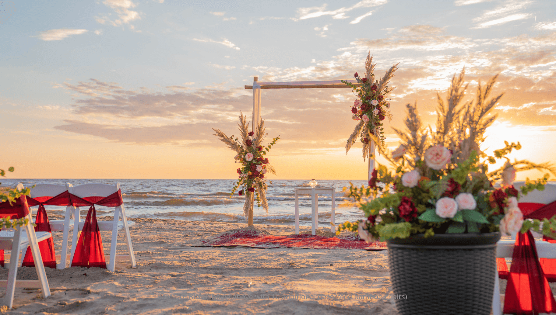 A beautifully decorated beach wedding setup at sunset featuring floral arrangements and chairs adorned with red sashes.