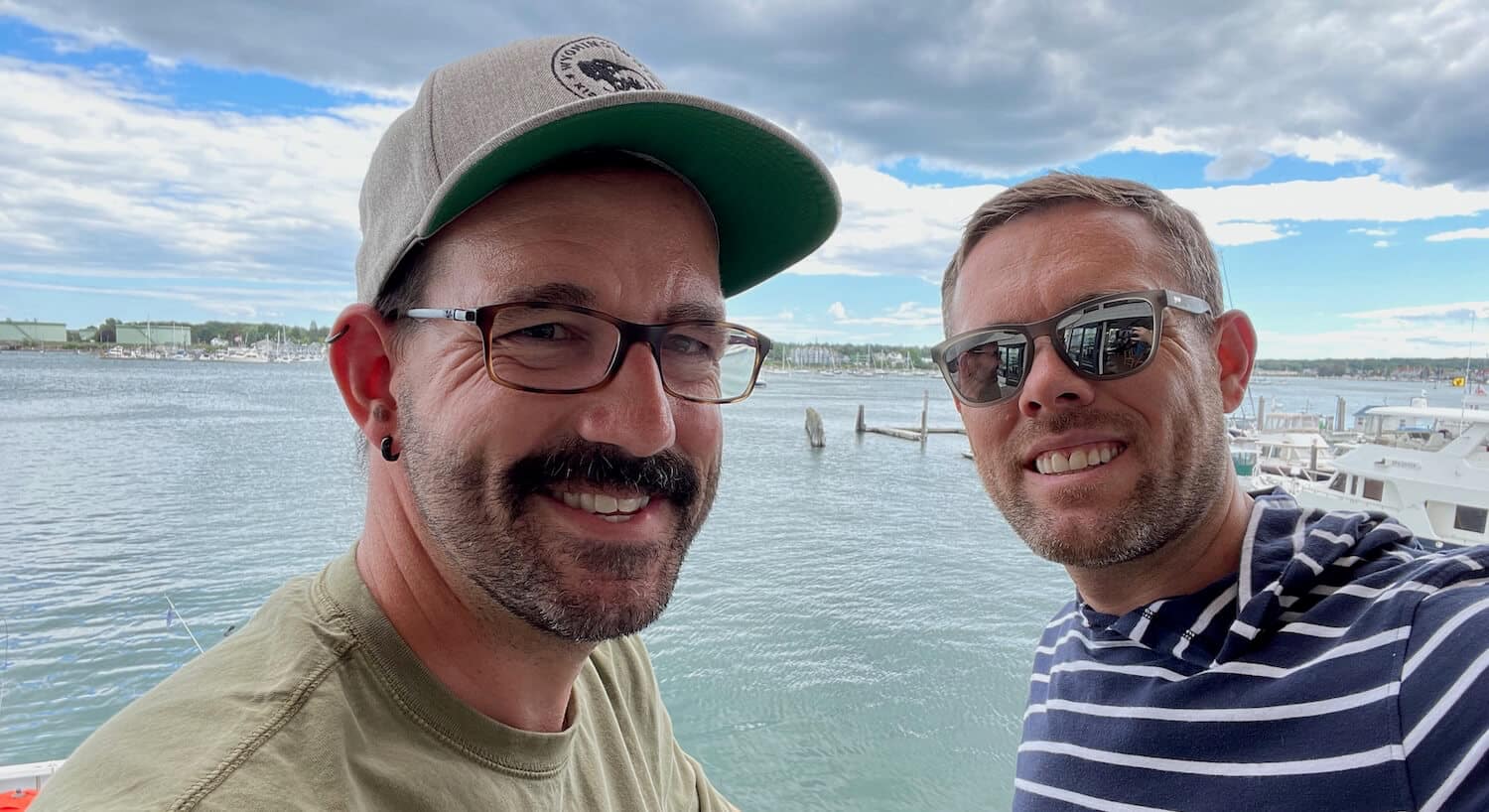 Two men smiling for a selfie with a marina and cloudy sky in the background.