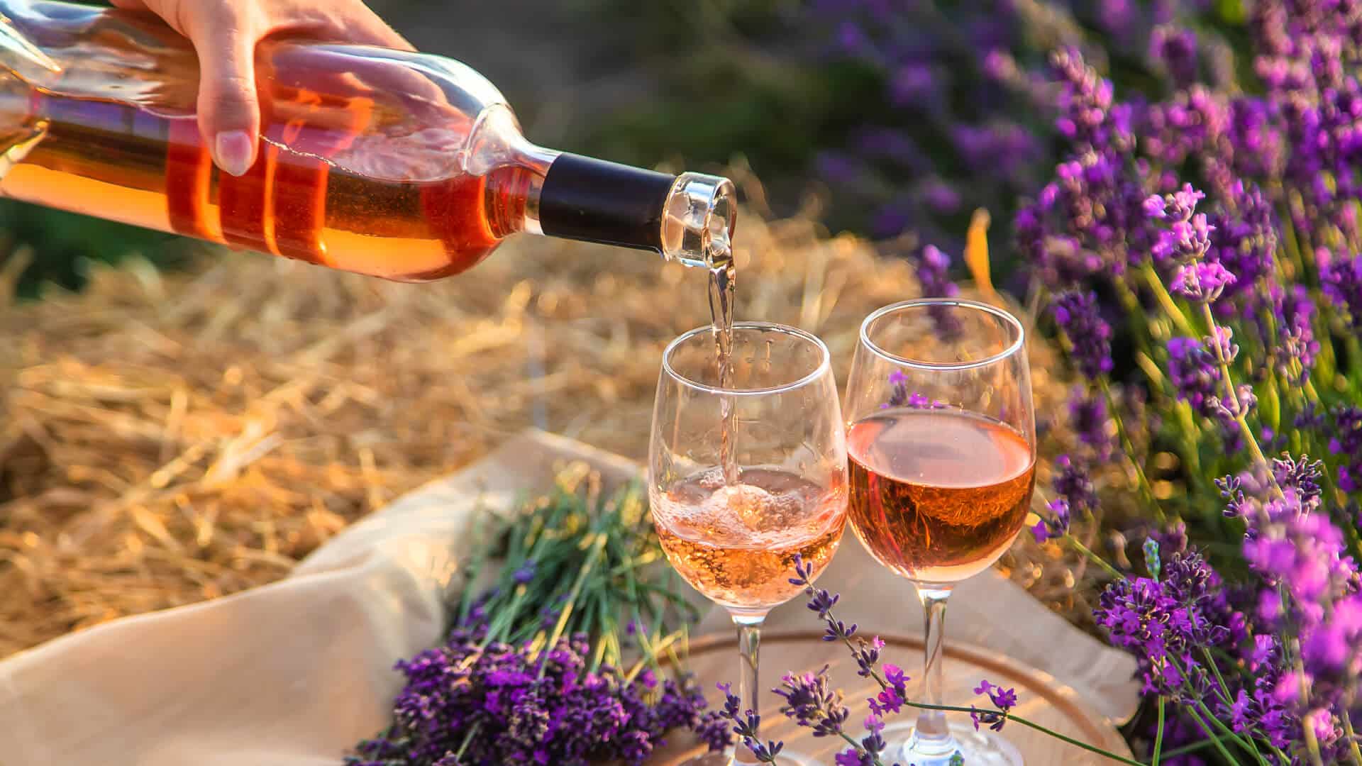 A hand pours rosé wine into two glasses surrounded by lavender flowers.