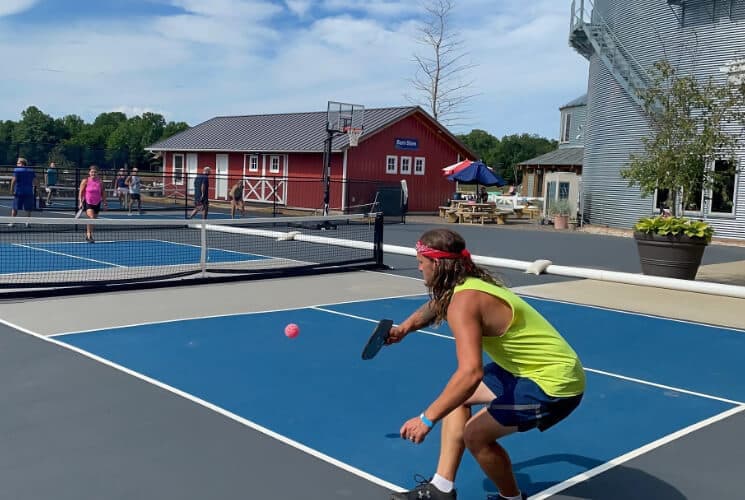 A player in a neon tank top prepares to hit a pink ball on a pickleball court with spectators in the background.