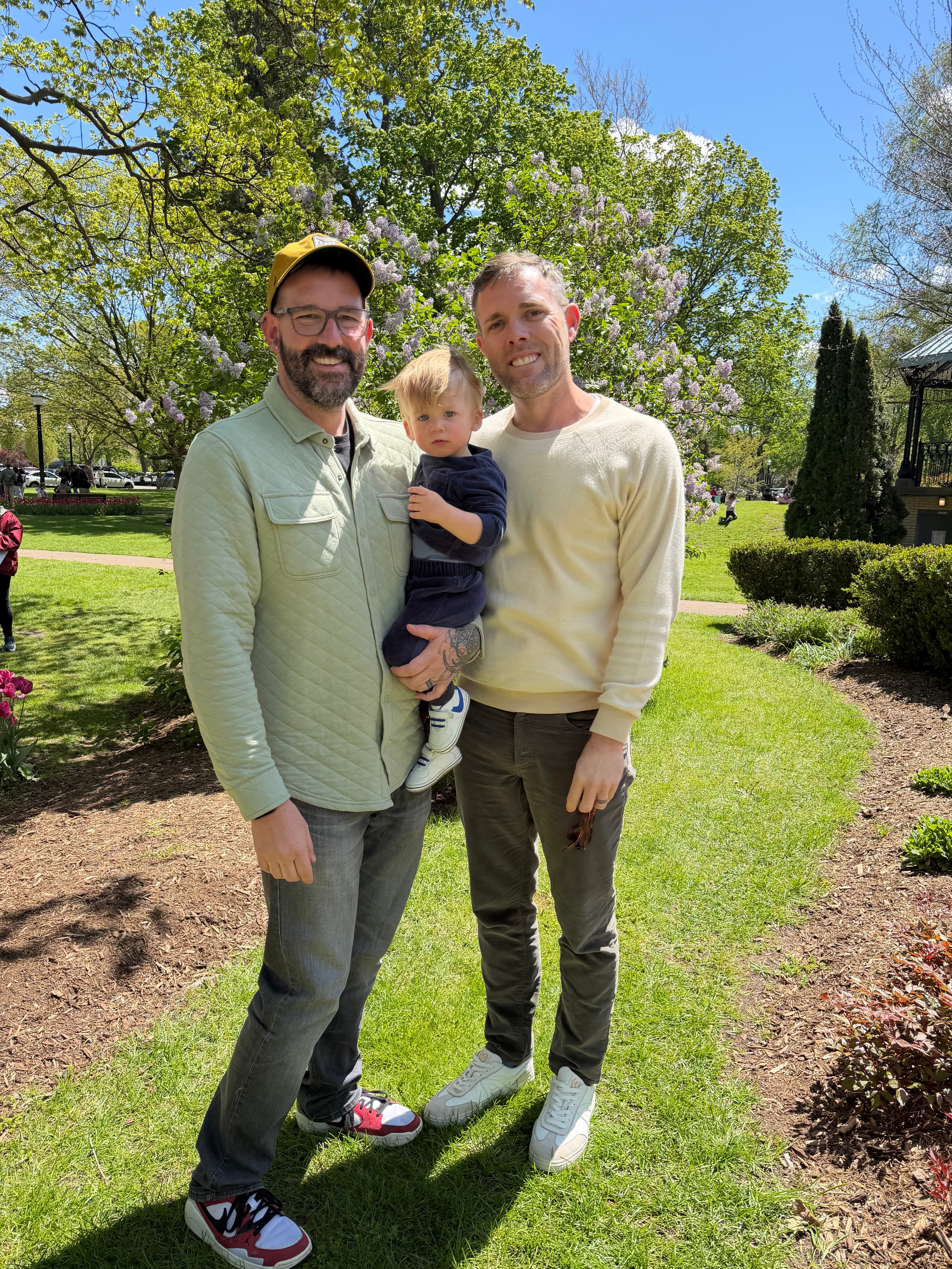 Two men smile while holding a young child in a sunny park.