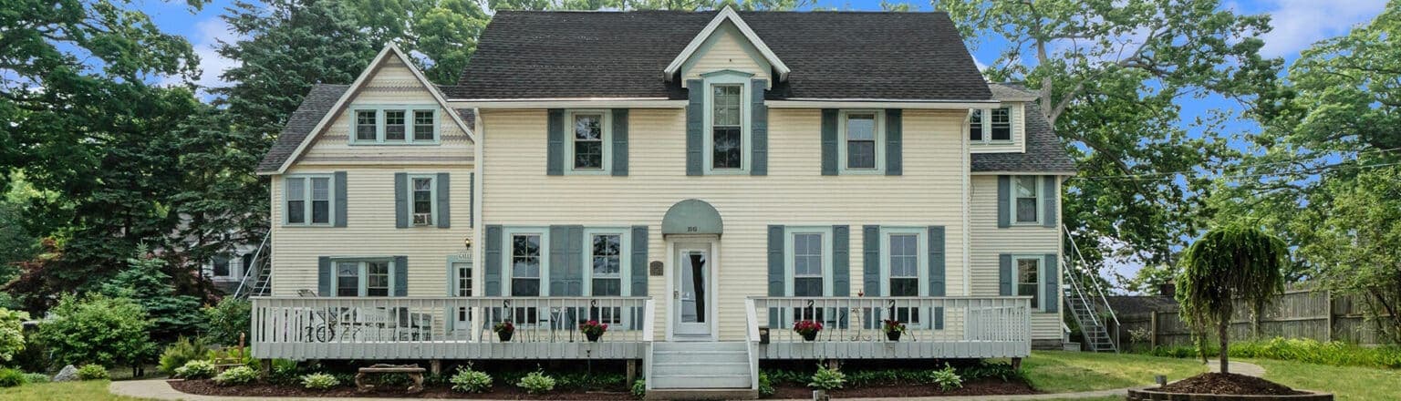 A large, two-story yellow house with green shutters and a welcoming front porch surrounded by greenery.