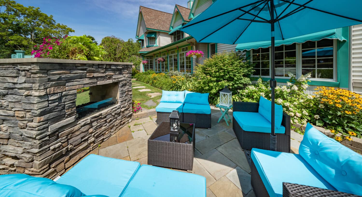 An outside patio area with wicker furniture with bright blue cushions and umbrella, facing a brick fireplace, green grass, bushes, and lots of flowers throughout.