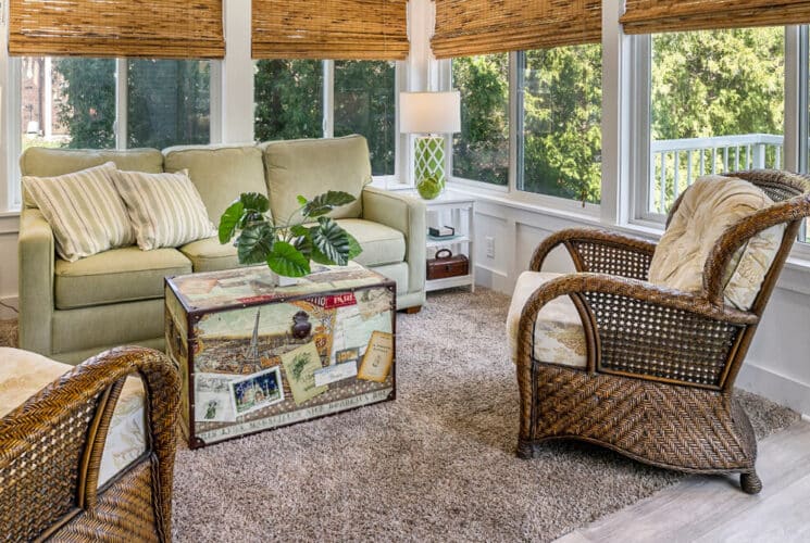 A sitting area with wood floors, a carpeted area rug, a sage green sofa, two wicker armchairs, a trunk as a coffee table, 2 side tables with lamps and windows with bamboo blinds, and views of the outdoor grounds and trees.