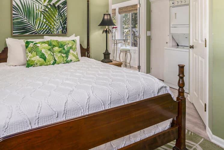 A bedroom with sage walls, white trim, a wood framed bed with white bedding with green leaves accent pillows, wicker nightstands with lamps on either side of the bed, a stackable washer and dryer.