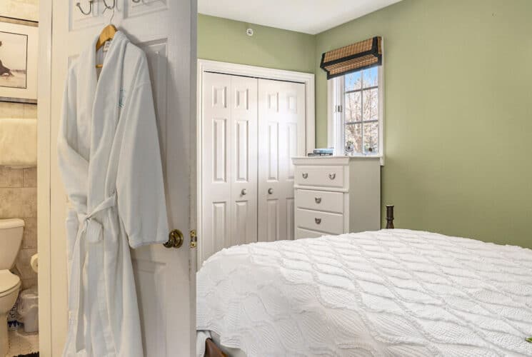 A bedroom with sage colored walls, a white chest of drawers, white doors, white bedding with green throw pillows, and a private bathroom with a robe hanging on the door.