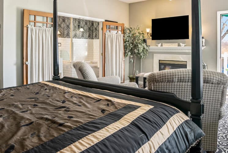 A bedroom with black and white carpeting, a canopy bed with black, brown and tan bedding, 2 plush armchairs, a fireplace, flat screen TV, a corner tub, and an open doorway leading to a balcony with a table.