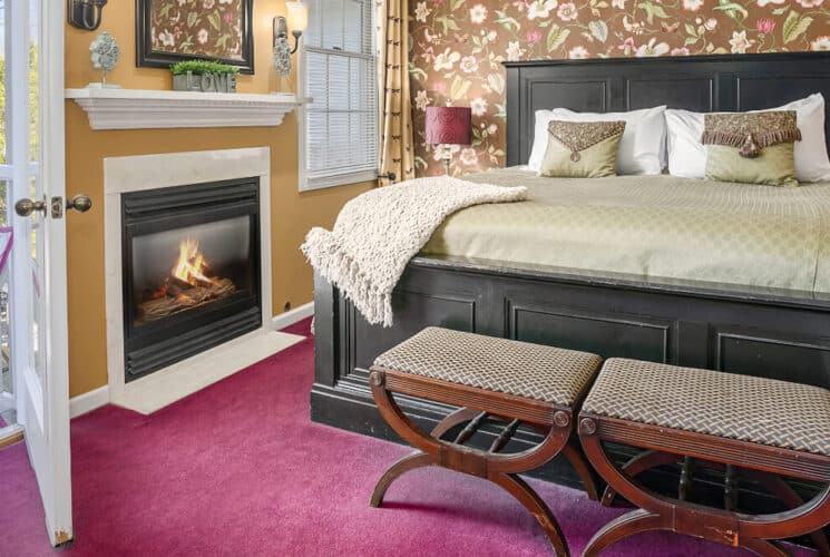 A bedroom with rose carpeting, a wood framed bed with tan and white bedding, 2 antique sitting chairs at the foot of the bed, a fireplace, and an open doorway to a private balcony with a metal table and chair.
