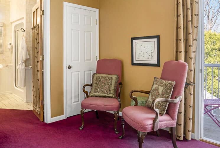 A room with rose carpeting, 2 pink armchairs, a writing desk, a private bathroom, and an open doorway leading to a terrace with a metal table and chairs.