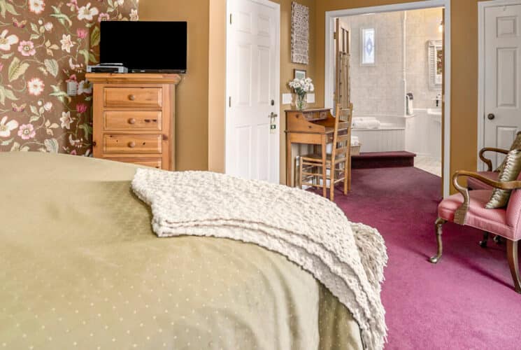 A bedroom with rose carpeting, brown floral wallpaper, a bed with tan bedding, a tall chest of drawers in the corner with a TV on top, a writing desk and a doorway leading to a private bathroom.