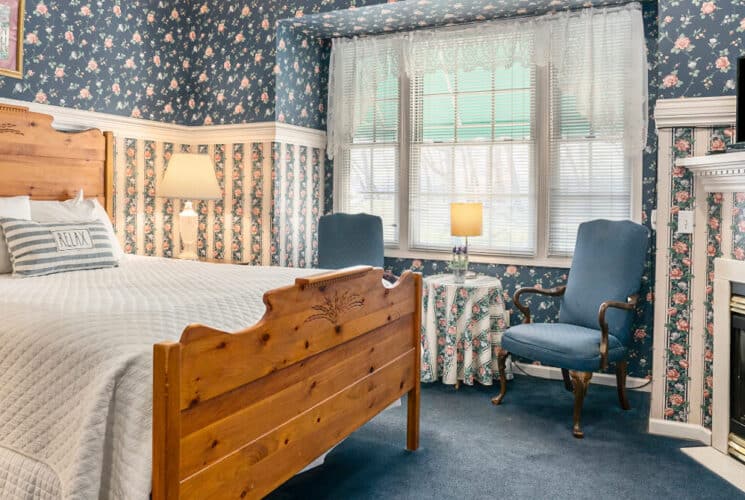 A bedroom with blue carpeting, floral striped wallpaper, and a wood framed bed with white bedding, a sitting area with blue chairs and a small table, along with a fireplace and flat screen TV on the mantle.