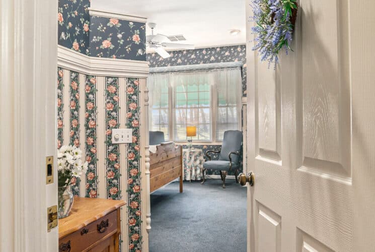 An open door leading to a bedroom with floral striped wallpaper, blue carpeting, a wood bed frame, and blue chairs and a table near a bank of 3 windows.
