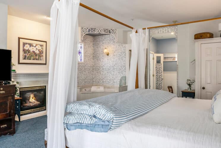 A bedroom with a canopy bed with white and blue bedding, an antique chest of drawers with a TV on top, a fireplace, and a corner soaking tub.