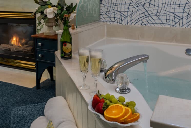 A bedroom with blue carpeting, a fireplace, and a corner soaking tub with fruit and champagne.