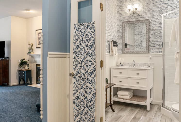 A bedroom with blue carpeting, an antique chest of drawers, a fireplace, and a private bathroom with a wood vanity with sink and corner shower.