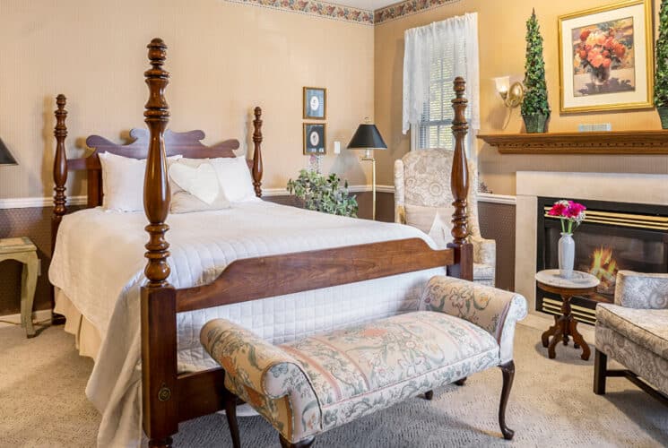 A bedroom with a four poster bed, a tapestry sitting bench at the foot of the bed, a sitting area with plush wingback chairs, and a fireplace.