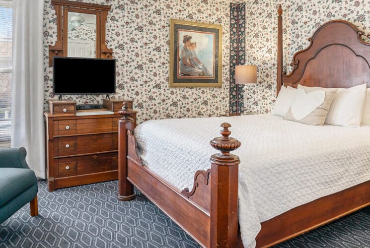 A bedroom with blue and white carpeting, an anitque wood framed bed with whit3e bedding, a nightstand with lamps on each side, an antique wood dresser with a TV and DVD player on it, a sitting area with blue wingback chairs, and floral wallpaper.