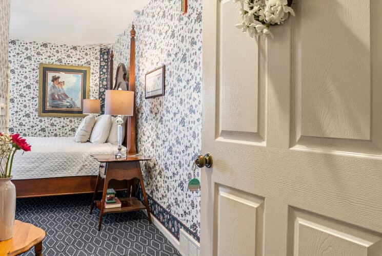 An open doorway leading to a bedroom with a wood framed bed with white bedding, blue and white carpeting, and floral wallpaper.