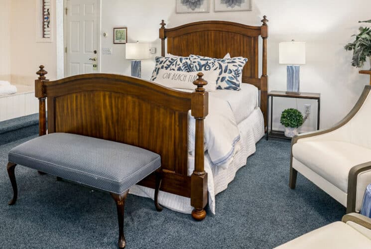 A bedroom with blue carpet, a wood framed bed with white and blue bedding, a blue sitting bench at the foot of the bed, a soaking tub built into a nook in the bedroom, and a sitting area with 2 plush chairs.