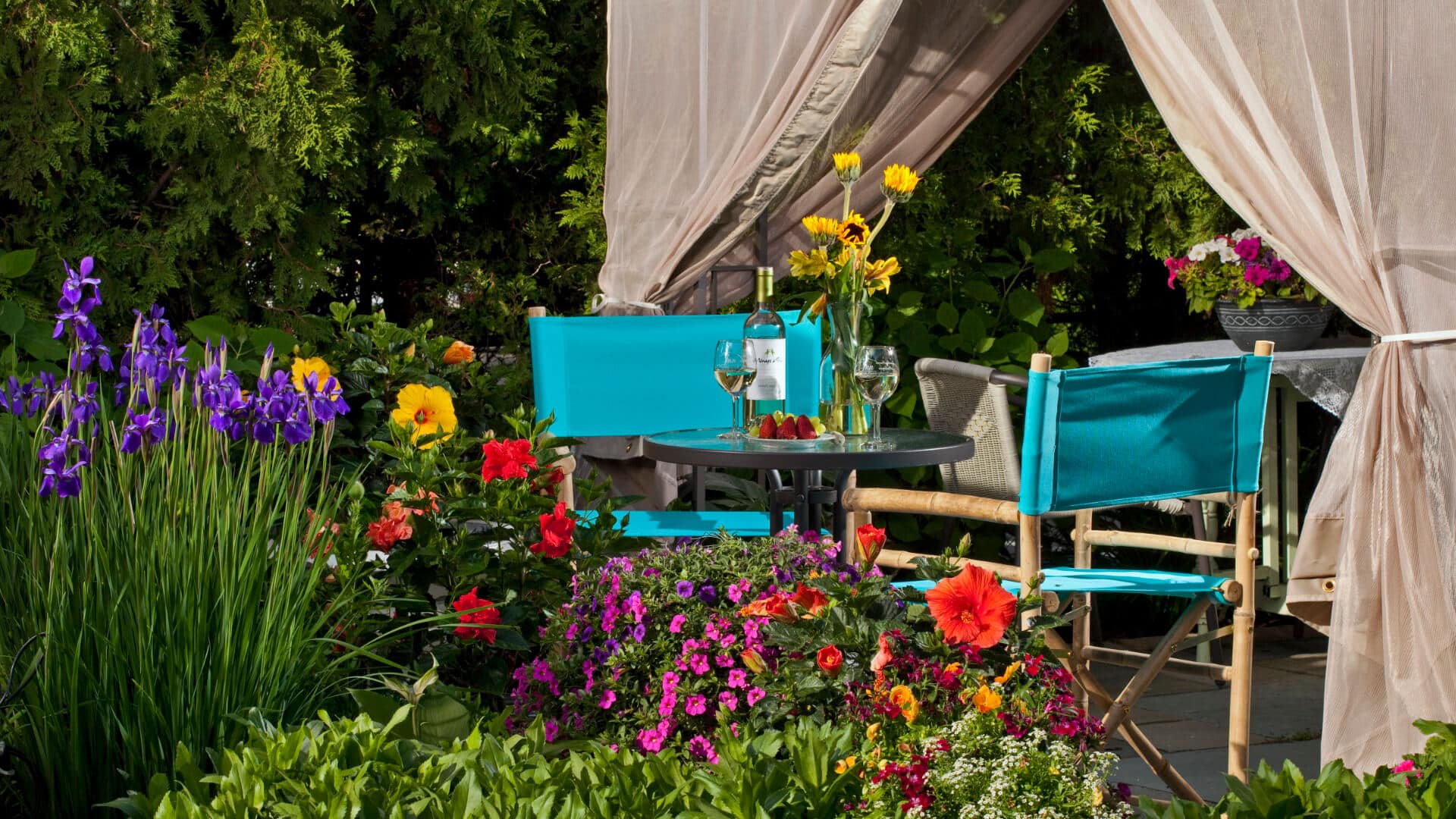 Round Glass table in garden with yellow, red and purple flowers and greenery, two white wine filled glasses, aqua chairs.