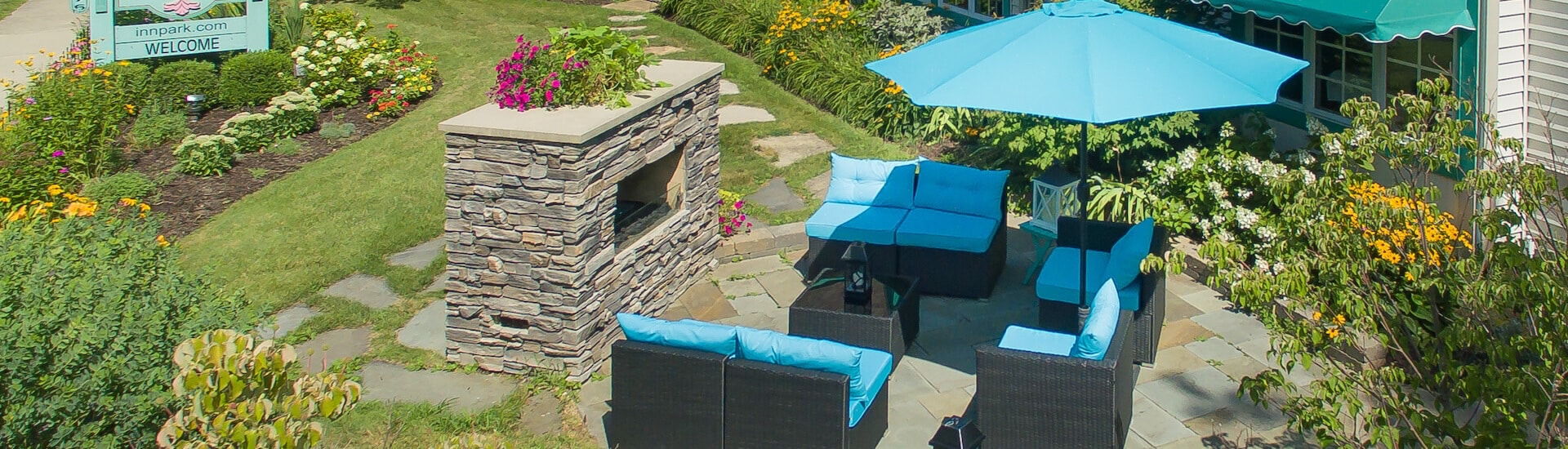 An outside patio area with wicker furniture with bright blue cushions and umbrella, facing a brick fireplace, green grass, bushes, and lots of flowers throughout.
