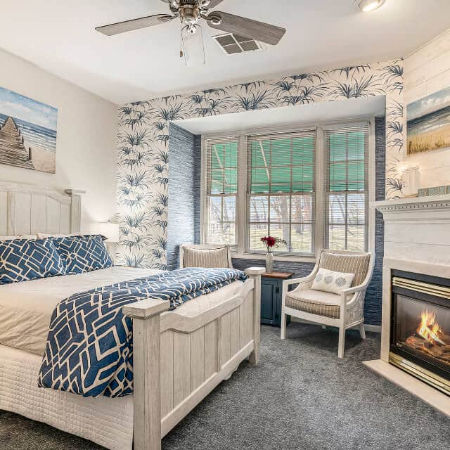 A bedroom with a white washed wood bed with blue and white bedding, a fireplace on the opposite wall, and a sitting area in a bay window.