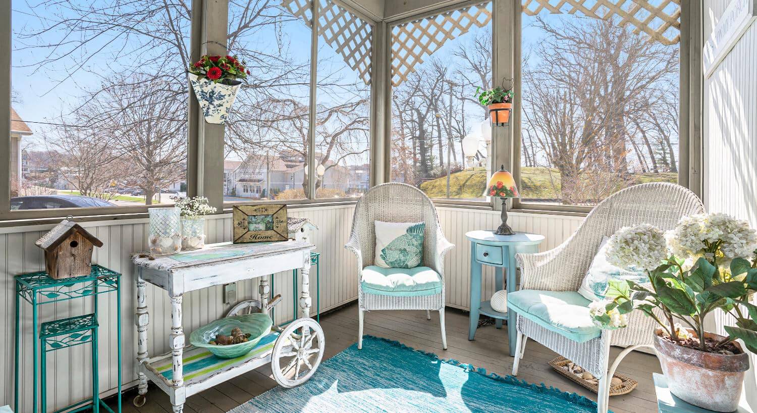 A screened in porch with white wicker furniture, wood floors, a blue area rug, along with pots of flowers.