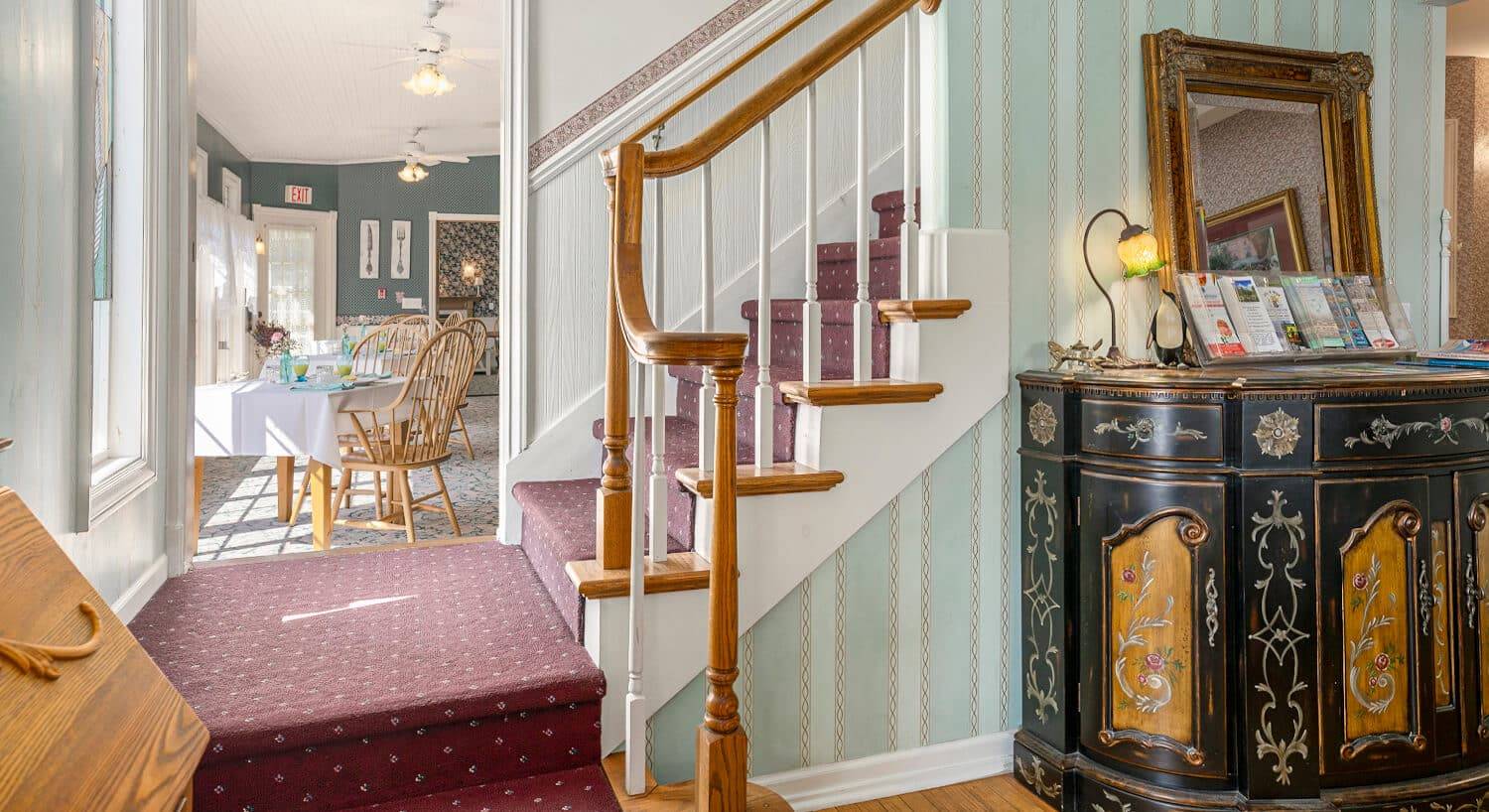 A staircase going up to a second floor, and 2 rooms on either side, one a dining room with tables and chairs, and the other a living area with antique furnishings.