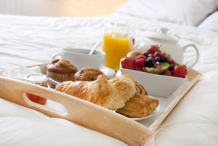 A tray on a bed with a variety of breakfast foods such a croissant, pastries, muffins, fresh fruit, jam, a glass of orange juice, a teapot and cup and saucer.
