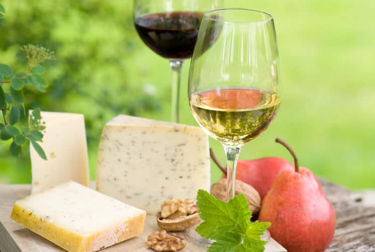 A wood board with a variety of cheeses, nuts, and fruit, with 2 glasses of wine, one red and one white.